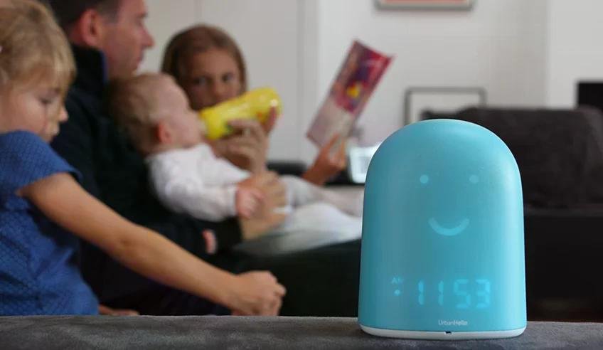 A blue, smiley-shaped baby monitor with a digital screen sits on a table while a family reads and feeds a baby in the