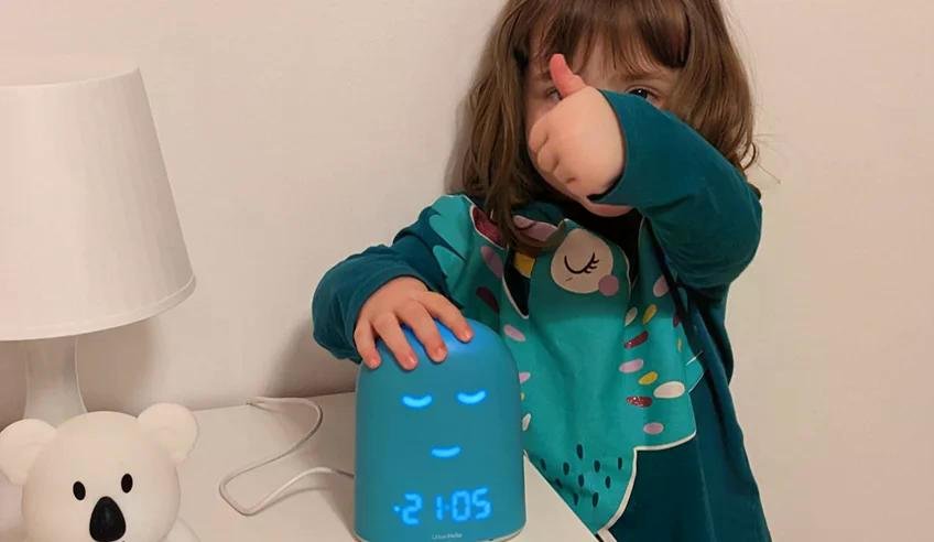 A child wearing a teal pajama top sits next to a blue digital clock with a sleeping face icon displayed.